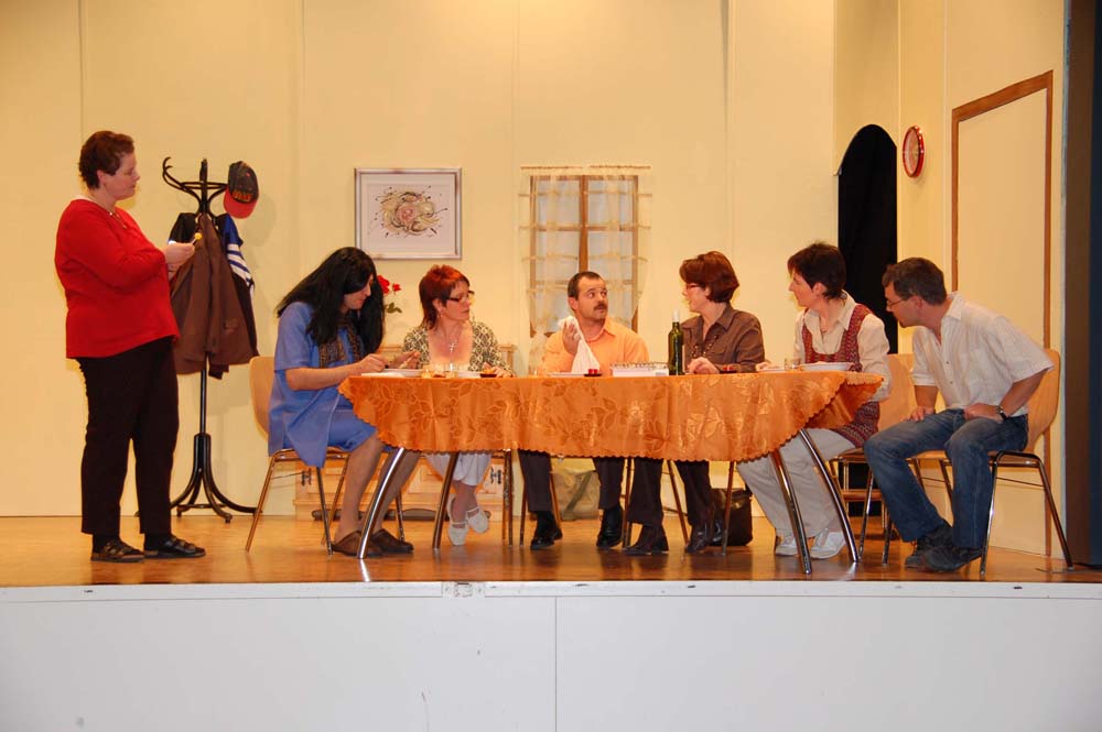 Sechs Theaterleute sitzen am Tisch auf der Bühne. Links daneben steht Monika.