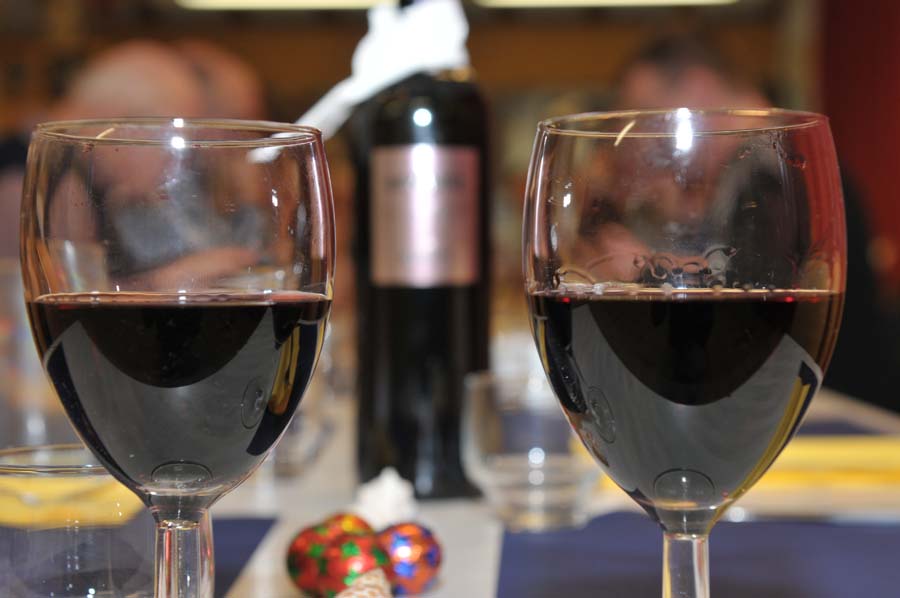 Zwei gefüllte Rotweingläser. Dazwischen ist im Hintergrund die dazugehörende Flasche zu sehen. 