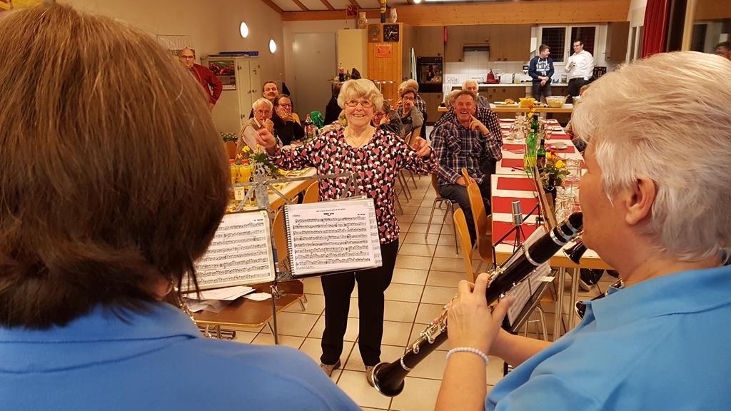 Die Jubilarin dirigiert einen Marsch. Links und rechts zwei Rücken von Holzbläserinnen.