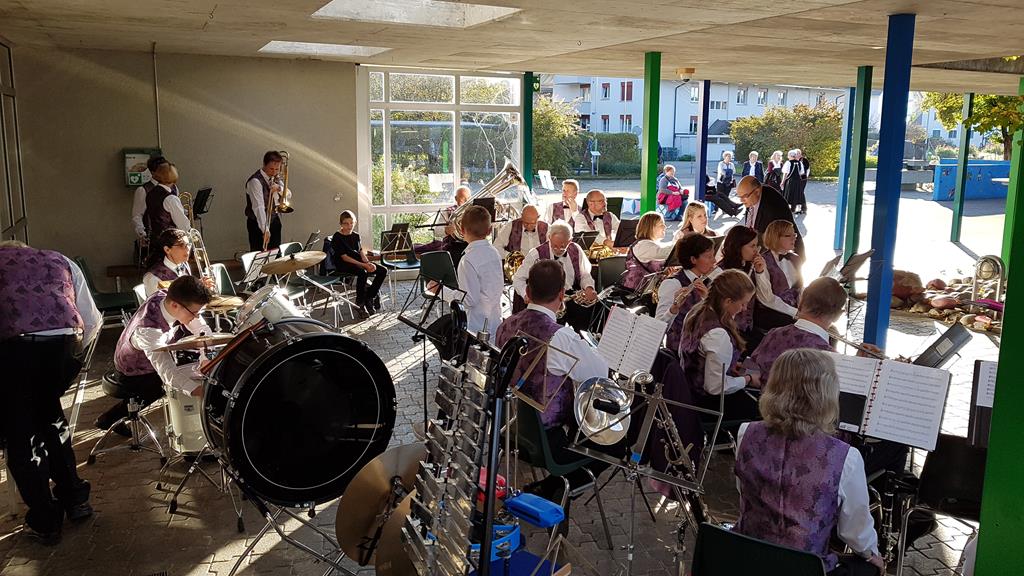 Die MGU spielt sitzend unter dem Baldachin beim Mehrzweckgebäude bei sonnigem Wetter